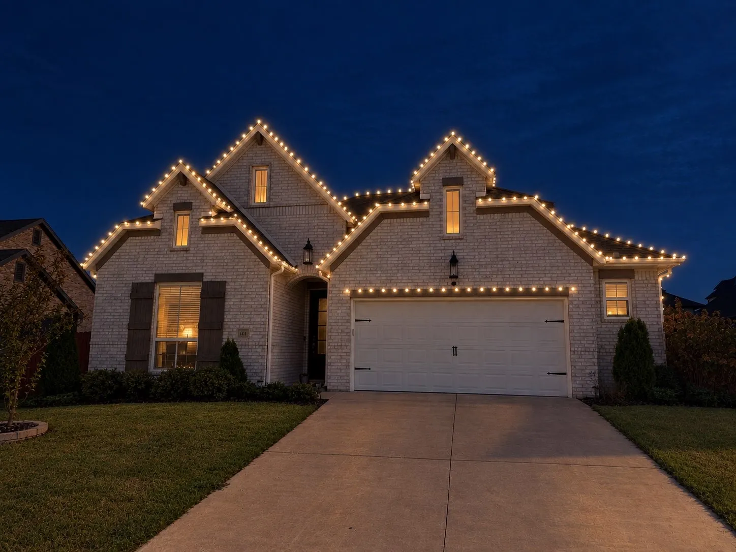 Two-story brick home with warm white lights tracing every roof peak and the garage line